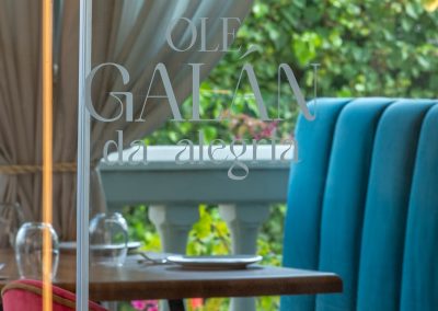 The glass facade of the Ole Galán da Alegría restaurant, showing the etched logo on the window. Inside, there is a dining table with a blue velvet booth and a pink velvet chair.